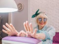 Manicure master in blue gloves creaming hands of elderly stylish woman in blue sunglasses and jeans jacket sitting at manicure sal