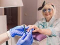 Manicure master in blue gloves creaming hands of elderly stylish woman in blue sunglasses and jeans jacket sitting at manicure sal