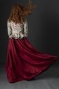 A mannequin with long curly red hair wearing a renaissance-style bodice and red skirt against a studio backdrop