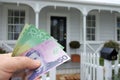 A mans hand holds NZ dollar bills against a front of North American hous