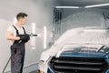 Manual car washing concept. Young man worker cleaning modern blue car with foam and pressured water at service station Royalty Free Stock Photo