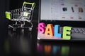 Many-colored wooden word SALE set on laptop bottom at the background of opened web page on the screen and small shopping trolley.