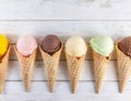 Many flavors and colors of scooped ice cream in waffle cones. Pistachio, vanilla, fruit, chocolate. White wooden background.