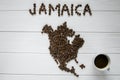 Map of the Jamaica made of roasted coffee beans laying on white wooden textured background with cup of coffee