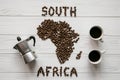 Map of the South Africa made of roasted coffee beans laying on white wooden textured background with coffe maker, cups of coffee