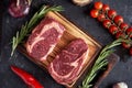 Marble ribeye steak on a cutting board with rosemary, peas, onions, garlic, red tomatoes on a branch on a concrete dark background