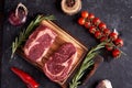 Marble ribeye steak on a cutting board with rosemary, peas, onions, garlic, red tomatoes on a branch on a concrete dark background