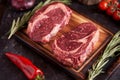 Marble ribeye steak on a cutting board with rosemary, peas, onions, garlic, red tomatoes on a branch on a concrete dark background