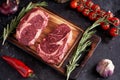 Marble ribeye steak on a cutting board with rosemary, peas, onions, garlic, red tomatoes on a branch on a concrete dark background