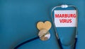 Marburg virus symbol. White card with words Marburg virus, beautiful blue background, wooden heart and stethoscope. Medical,