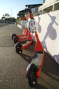 Four orange and black Neuron rent to ride electric scooters with white protective helmets on a pavement sidewalk beside Auckland H