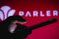 March 1, 2021, Brazil. In this photo illustration the Parler logo seen in the background of a silhouette hand holding a mobile