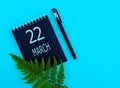 March 22th. Day 22 of month, Calendar date. Black notepad sheet, pen, fern twig, on a blue background