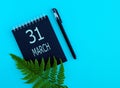 March 31th. Day 31 of month, Calendar date. Black notepad sheet, pen, fern twig, on a blue background