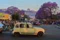 Market day in Antananarivo, a vintage renault taxi drives near jacaranda trees blossoming