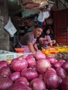 A market vendor sells red onions and other produce, surrounded by bustling shoppers and the enticing aroma of fresh vegetables.