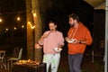 Married couple man and woman eating barbecue sausages at night near lightning garlands in the backyard