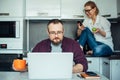 Married couple using laptop and smartphone in the kitchen, deep in their gadgets. Serious man with beard working online at home.