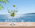 Martini glass with olives and beverage splash is on table top. Blurred sparkling sea at the background. Place for your product or Royalty Free Stock Photo