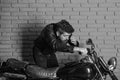 Masculinity concept. Man with beard, biker in leather jacket near motor bike in garage, brick wall background. Hipster