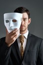 The mask can be misleading. Studio portrait of a young businessman holding a mask in front of his face against a gray