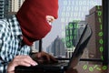 Masked hacker wearing a balaclava hacking data from laptop against digital city background