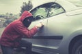 Masked man in casual wear furtively picks the car lock on a city background. Male thief stealthily picklocks the vehicle