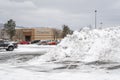 A massive pile of plowed snow after a winter storm sits in a parking lot of a shopping mall with a department store in view behind