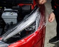 The master applies vinyl film to the headlight of a red car. Closeup view on worker detailer hand smoothing with a