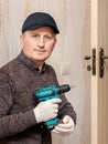 The master with a cordless screwdriver in his hands in the room near the door intends to repair the door lock