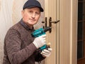 The master with a cordless screwdriver in his hands in the room near the door intends to repair the door lock