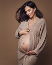 Maternity Knitwear. Pregnant Woman in Woolen Cardigan looking at Belly over Beige Background. Pregnancy Fashion Clothes. Maternal Royalty Free Stock Photo