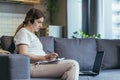 Maternity leave. Work. Pregnant freelancer woman sitting at home on sofa with laptop and in headphones in home clothes. Works on Royalty Free Stock Photo