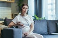 Maternity leave. Work. Pregnant freelancer woman sitting at home on sofa with laptop and in headphones in home clothes. Works on Royalty Free Stock Photo