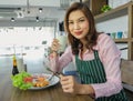 Matue adult Asian female housewife in apron sitting at table in kitchen holding glass of milk and ready to eat breakfast. Happy