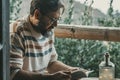 Mature adult man sitting at home in the outdoor terrace reading a book and relaxing enjoying the nature outside in background - Royalty Free Stock Photo