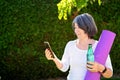 Mature adult woman practicing fitness outdoors holding bottle of water and yoga mat looking at online guide at smartphone. Senior Royalty Free Stock Photo