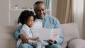 Mature african american man with cute kid girl hugging sitting in room on sofa father reads greeting card loving