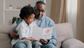 Mature african american man with cute kid girl hugging sitting in room on sofa father reads greeting card loving