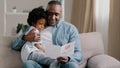 Mature african american man with cute kid girl hugging sitting in room on sofa father reads greeting card loving