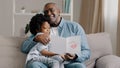 Mature african american man with cute kid girl hugging sitting in room on sofa father reads greeting card loving