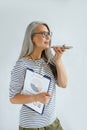 Mature Asian businesswoman records audio message on cellphone holding diagrams in studio