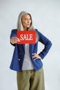 Mature Asian lady with grey hair in jacket holds red card with word Sale in studio