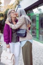 Mature Asian woman hugs and kisses positive friend meeting on modern city street