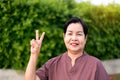 Mature asian woman standing and showing 2 fingers at public park,Happy and smiling,Senior care insurance concept