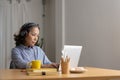 Mature asian woman wearing headphones using laptop, making video call, sitting at table in kitchen, senior teacher Royalty Free Stock Photo