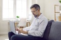 Mature man doing remote work working on laptop in his home office making notes sitting on couch. Royalty Free Stock Photo