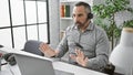 A mature bearded man with grey hair engages in a video call at his office workspace, exuding professionalism