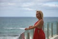 Mature, beautiful, blonde woman in a red dress, leaning on a glass railing looking out to sea while the wind stirs her hair. Royalty Free Stock Photo