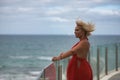 Mature, beautiful, blonde woman in a red dress, leaning on a glass railing looking out to sea while the wind stirs her hair. Royalty Free Stock Photo